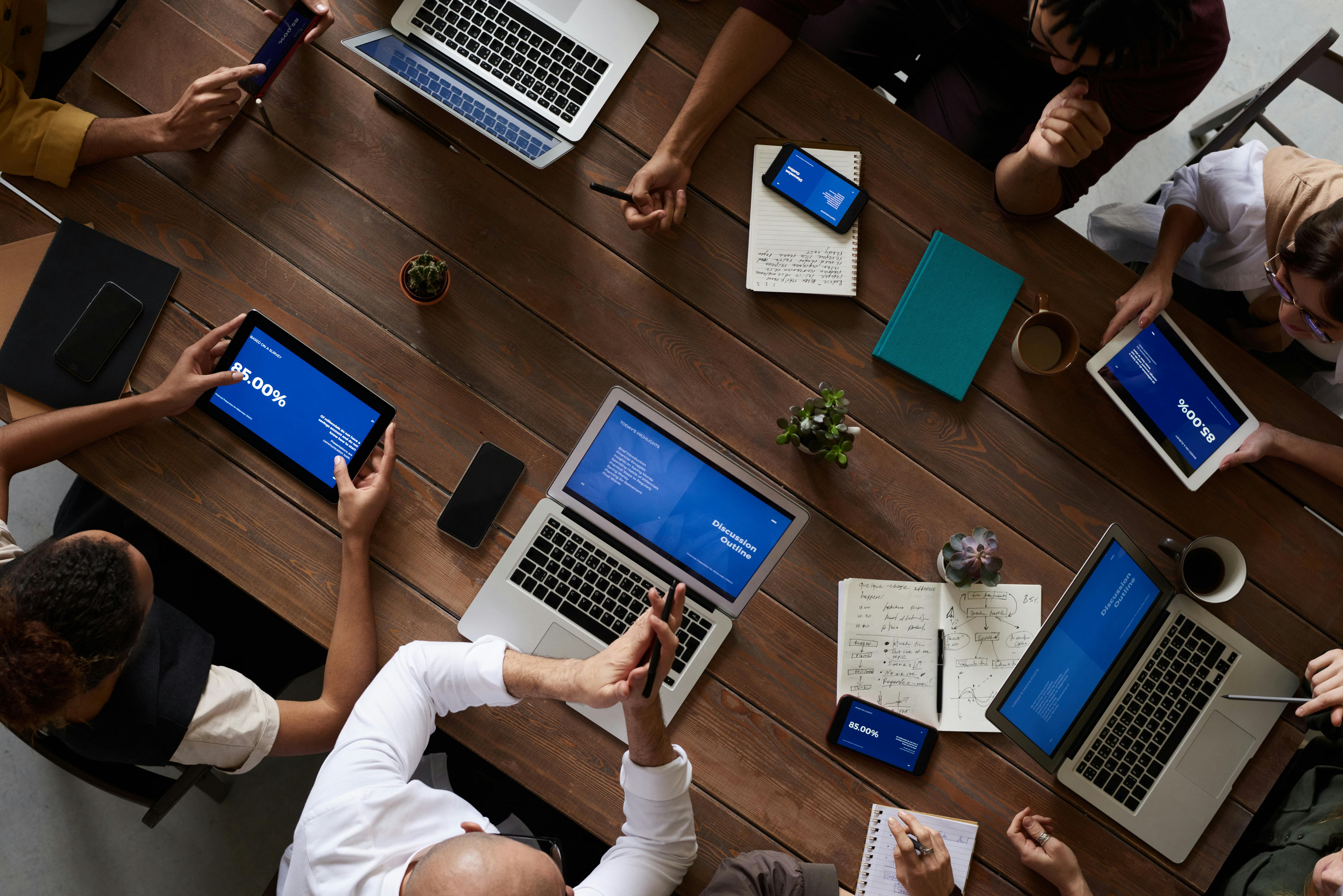 Team collaborating around a wooden table with laptops, tablets, notebooks, and digital marketing presentations on screens